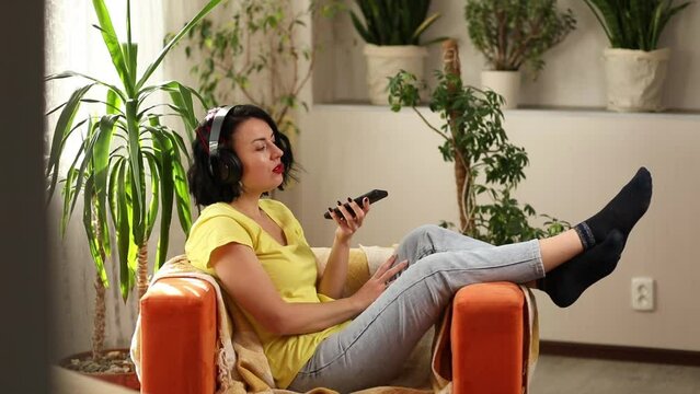 Cheerful woman in casual clothes with headphones and smartphone, record to podcast on sofa at home. Female podcaster live streaming, Woman speaking in clubhouse.
