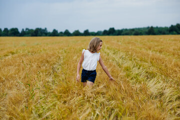 Portrait of a girl in a wheat field. She walks and strokes the ears with her hands. Summer rural landscape.