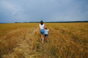 Naklejka na ściany i meble A happy family of mother and son in a summer wheat field against a stormy sky. They run across the field with their backs to the camera. Rural landscape.