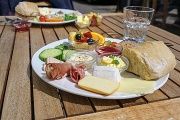 Varied breakfast or lunch plate in a street restaurant with sausage and ham, bread rolls, cheese and fruits on a wooden outdoor table on a sunny day, selected focus
