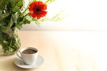 Coffee cup and a flower bouquet in a glass vase on a light wooden table, large copy space, selected focus