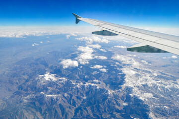 The wing of an airplane flying over clouds and mountains. Aerial view.