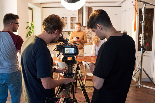Rear view of videographer and his assistant adjusting video camera for shooting of commercial or cooking masterclass in the kitchen