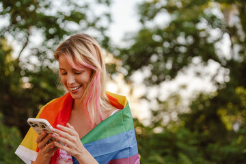 Young lesbian girl wrapped in rainbow flag holding smartphone outdoors