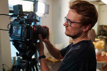 Young videographer or cameraman adjusting focus of his video camera while standing in studio and preparing for shooting advertisement