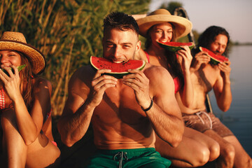Group of young people eating watermelon on a dock by the river during the summer sunny day