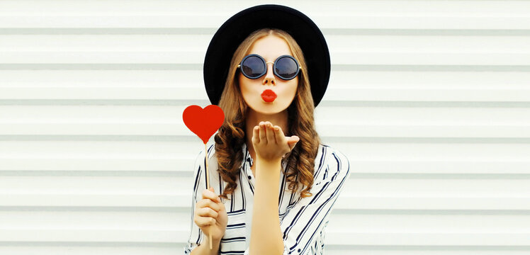 Portrait of stylish young woman blowing her red lips sending air kiss with red heart shaped lollipop wearing black round hat, striped shirt on white background