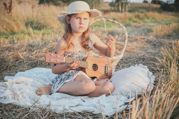 Portrait of girl sitting on blanket in dry hay field, having picnic, learning playing guitar...