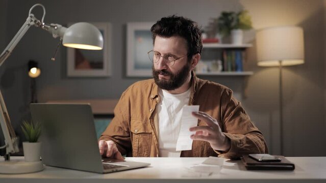 Positive man enters data from pay check into computer. Beard freelancer in home office enters data into app and calculates expenses. Laptop user doing home bookkeeping in laptop app