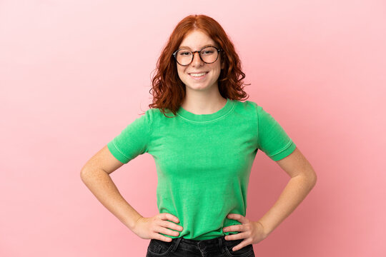 Teenager redhead girl over isolated pink background posing with arms at hip and smiling