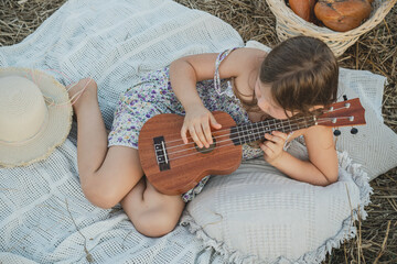 Portrait of girl lying on blanket in dry hay field, having picnic, learning playing guitar ukulele....