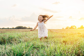 Portrait of smiling girl playing, jumping and running on grass hay field paths of dry grass in the sunset. Waving hands. Forest on bright light background. Cloudy sunny sky. Haying time - Powered by Adobe
