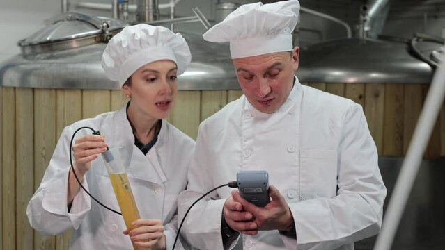 Interested focused male and female brewers wearing white uniform controlling process of craft beer production in small brewery, measuring pH level with digital meter. High quality 4k footage
