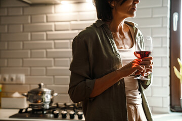 Mature brunette woman drinking wine while standing in kitchen