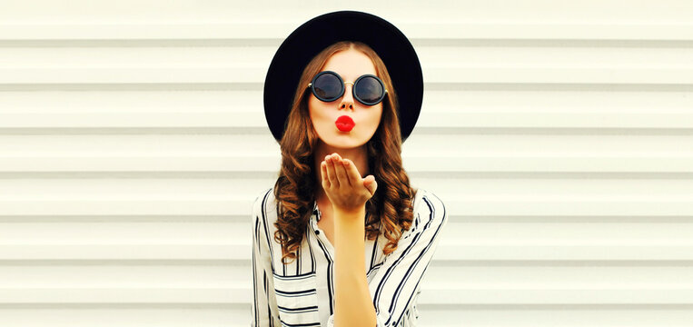 Portrait of stylish young woman blowing her red lips sending air kiss wearing black round hat, striped shirt on white background