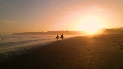 AERIAL SILHOUETTE: Following two surfers walking down the beach carrying surfboards. Surf friends checking waves in golden light after surf session. Gorgeous summer scenery at Playa Venao in Panama. - Powered by Adobe