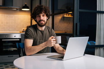 Fototapeta na wymiar Portrait of friendly bearded young freelancer male holding in hand cup with morning coffee sitting at table with laptop computer, looking at camera, in kitchen with modern interior.