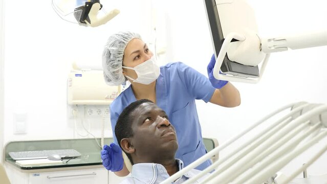 Asian woman dentist pointing at display to show information about teeth to african-american man patient during consultation.