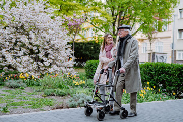 Senior man with walking frame and adult daughter outdoors on a walk in park.