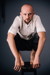 Portrait of a bald guy with a beautiful beard in a white shirt and plaid trousers squatting on a bar stool on a gray background