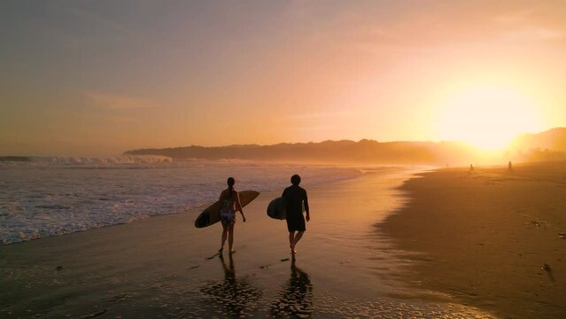 AERIAL SILHOUETTE: Two surfers carrying surfboards and checking waves after surf session. Surf friends walking on the beach in golden light. Gorgeous summer scenery at Playa Venao in Panama.