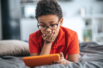 A curly-haired boy in orange tshirt looking involved while watching something online
