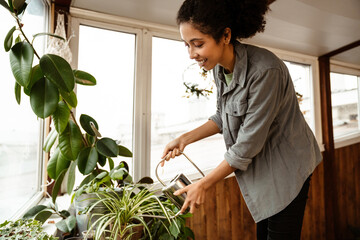 Young black woman wearing shirt watering plants in home