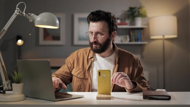 Man works at computer in home office. Freelancer working using laptop and smartphone. Bearded remote worker in glasses setting up streaming broadcast