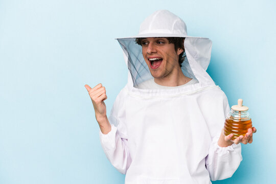 Young caucasian beekeeper man isolated on blue background points with thumb finger away, laughing and carefree.