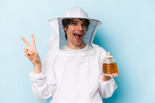 Young caucasian beekeeper man isolated on blue background joyful and carefree showing a peace symbol with fingers.