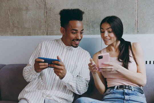 Two young friends woman man of African American ethnicity 20s in casual clothes hold in hand use show mobile cell phone play games sitting on gray sofa indoor spend free time rest in flat together