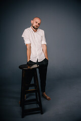 Portrait of a bald man with a beautiful beard in a white shirt and plaid pants near a bar stool on a black background