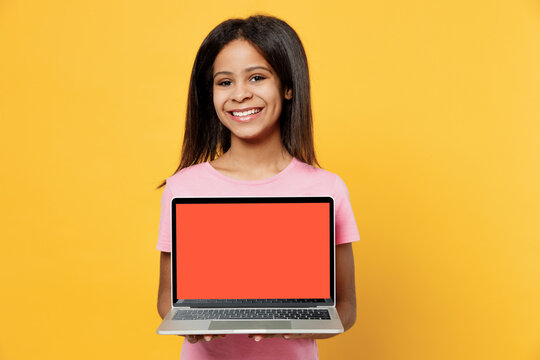 Little happy kid girl of African American ethnicity 12-13 years old wear pink t-shirt hold use work on laptop pc computer with blank screen workspace area isolated on plain yellow background studio