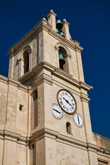 Church tower in Malta with multiple clocks on it in front of a clear dark blue sky on a sunny day....