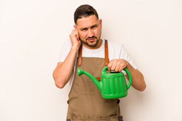Gardener hispanic man holding a watering can isolated on white background covering ears with hands. © Asier