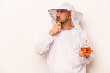 Hispanic beekeeper man holding honey isolated on white background looking sideways with doubtful and skeptical expression.