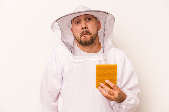 Hispanic beekeeper man holding honey isolated on white background shrugs shoulders and open eyes confused.