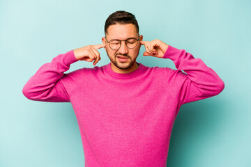 Young hispanic man isolated on blue background covering ears with fingers, stressed and desperate by a loudly ambient.