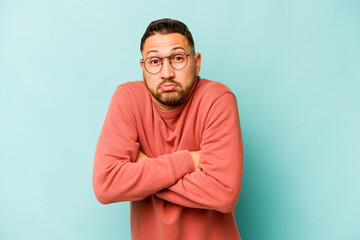 Young hispanic man isolated on blue background shrugs shoulders and open eyes confused.