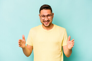 Fototapeta premium Young hispanic man isolated on blue background holding something with both hands, product presentation.