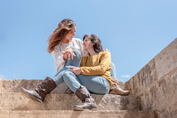 a lesbian couple embracing and looking each other in the eyes sitting on the stairs, in the background blue sky, concept of Lgbt, bisexuality, relationship lifestyle concept.