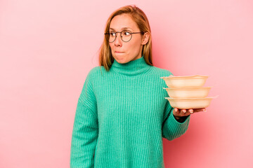 Young caucasian woman holding tupperware isolated on pink background confused, feels doubtful and unsure.