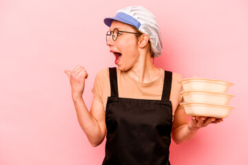 Young cook volunteer woman isolated on pink background points with thumb finger away, laughing and carefree.