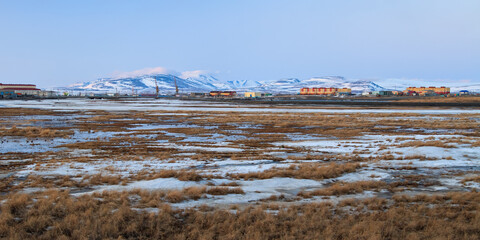 Spring arctic landscape. View of the tundra, colorful buildings and mountains. Beautiful evening panorama. Melting snow in the tundra on the river bank. Tavayvaam, Anadyr, Chukotka, Siberia, Russia.