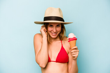 Young caucasian woman wearing a bikini and holding an ice cream isolated on blue background covering ears with hands.