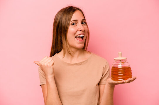 Young caucasian woman holding honey isolated on pink background points with thumb finger away, laughing and carefree.