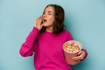 Young caucasian woman holding popcorn isolated on blue background shouting and holding palm near opened mouth.