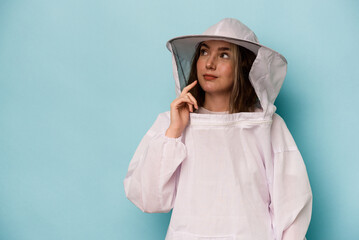 Young caucasian beekeeper woman isolated on blue background looking sideways with doubtful and skeptical expression.