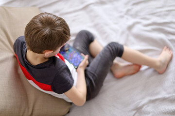 Little boy playing mobile game on smartphone or watching cartoons sitting on a sofa, top view. Child leisure at home, video gaming addiction