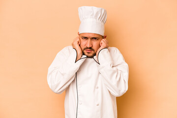 Hispanic chef man isolated on beige background covering ears with hands.